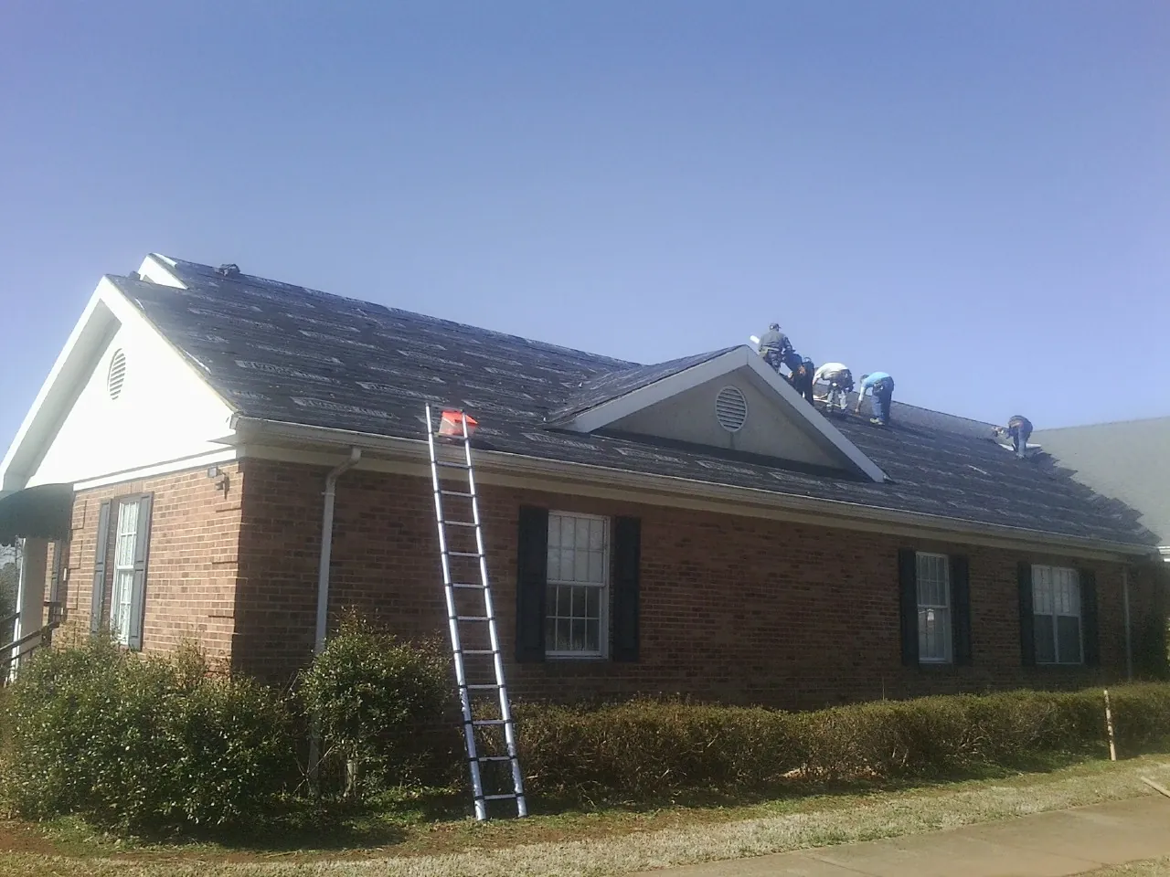 Crew tearing shingles off a brick ranch roof with a ladder set — Emergency Roof Repair work by our crew in Pennington
