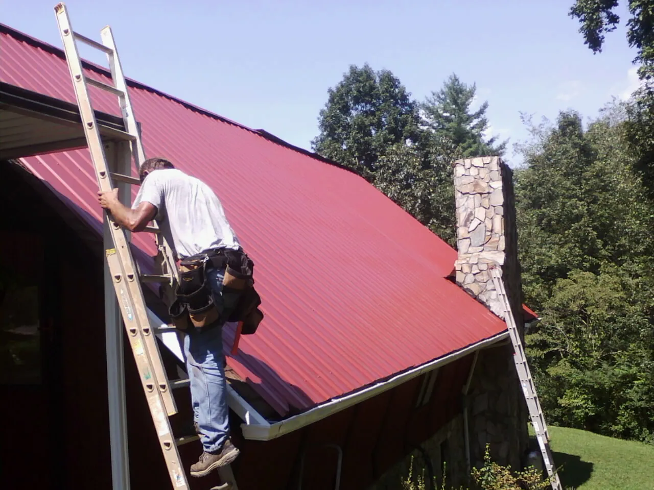 Roofer on a ladder beside a red metal roof and stone chimney — Soffit & Fascia Repair by Roofing Brothers in Pennington
